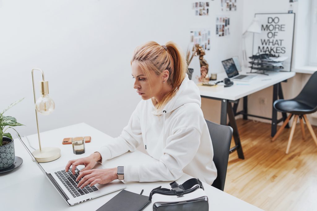 Peaceful and comfortable office and female worker in white hoody types on laptop sitting at table there is virtual reality headset.